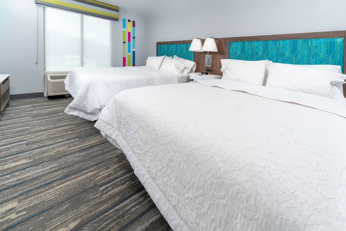 A hotel room with two beds, a window with blinds, a wall with colorful vertical stripes, and a patterned carpet.