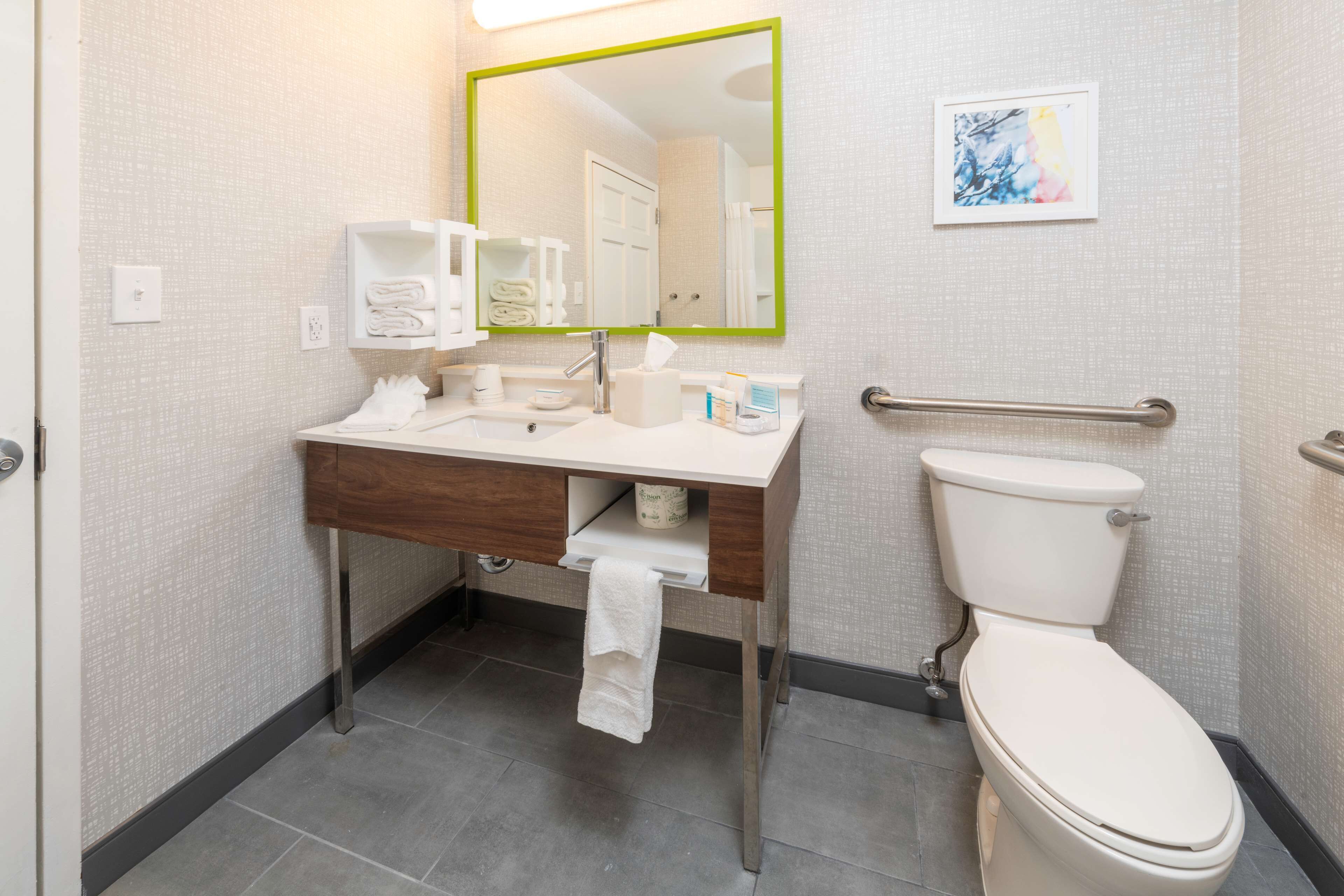 Une salle de bain avec un lavabo blanc, un miroir et des toilettes. On y trouve un porte-serviettes et une barre d’appui.