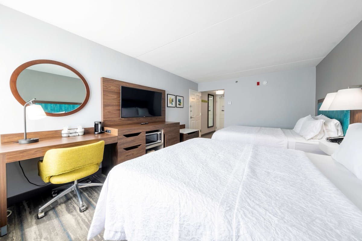 A hotel room with a bed, a desk with a chair, a TV, a mirror, and a lamp.