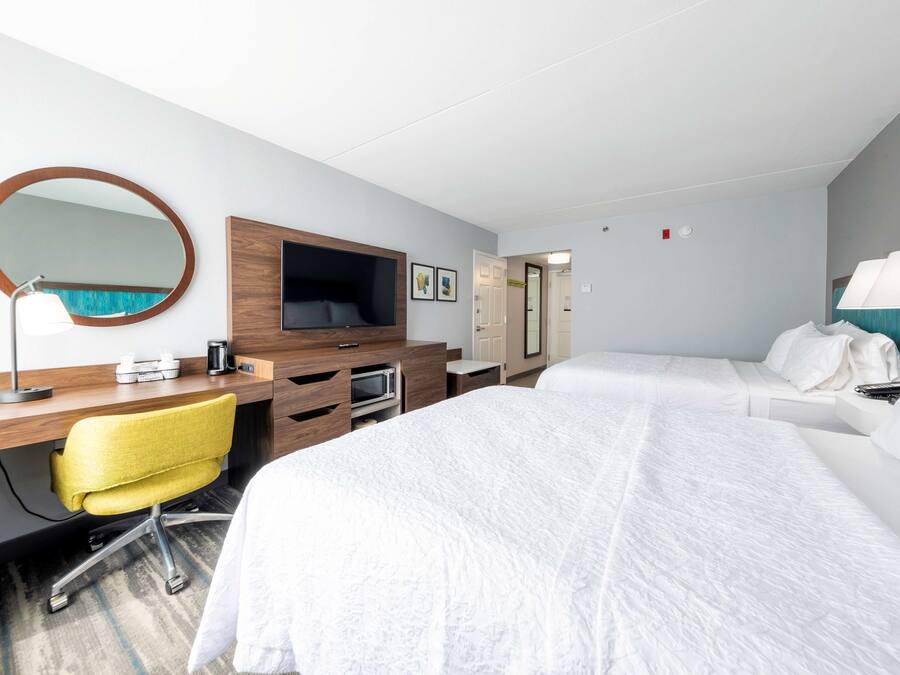 A hotel room with a bed, a desk with a chair, a TV, a mirror, and a lamp.