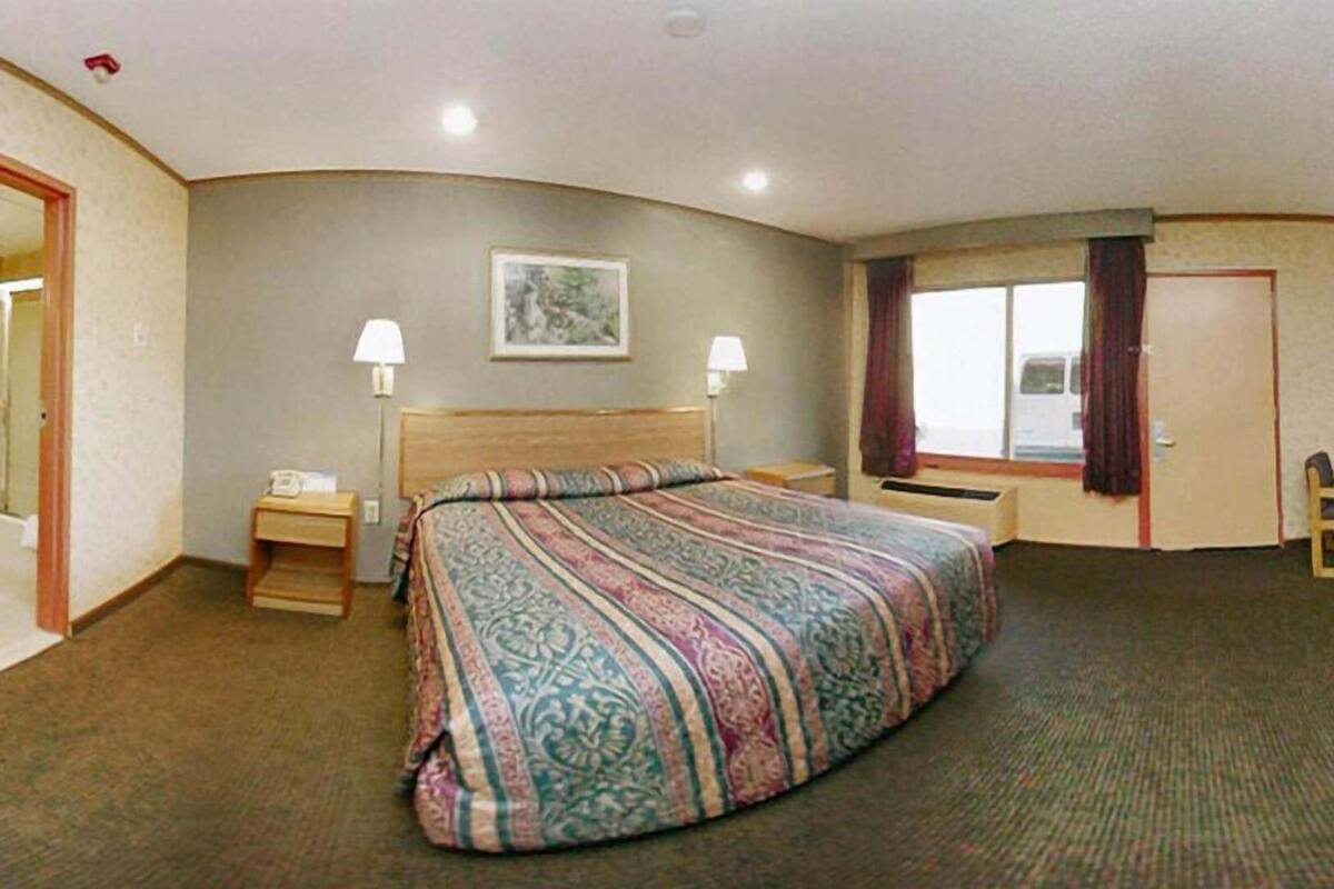 Hotellihuone, jossa on suuri sänky, kaksi yöpöytävalaisinta, ikkuna, jossa on verhot, pieni pöytä ja tuoli sekä ovi.