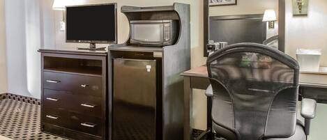 Een kleine hotelkamer met een flatscreen televisie, een magnetron, een kleine koelkast, een bureau met stoel en een spiegel in de badkamer.