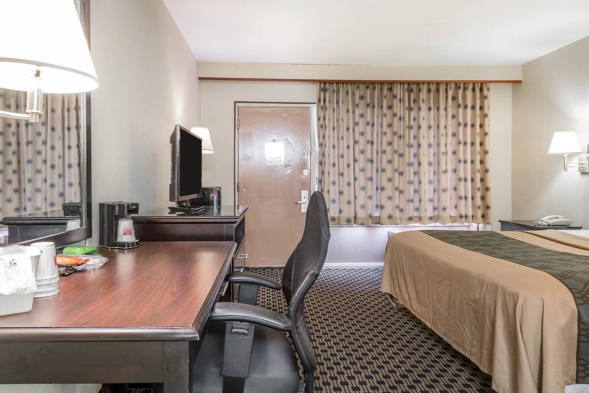 Ett hotellrum med ett skrivbord i trä, en tv, en säng med ett beige överkast, en mönstrad matta och en dörr med gardiner.