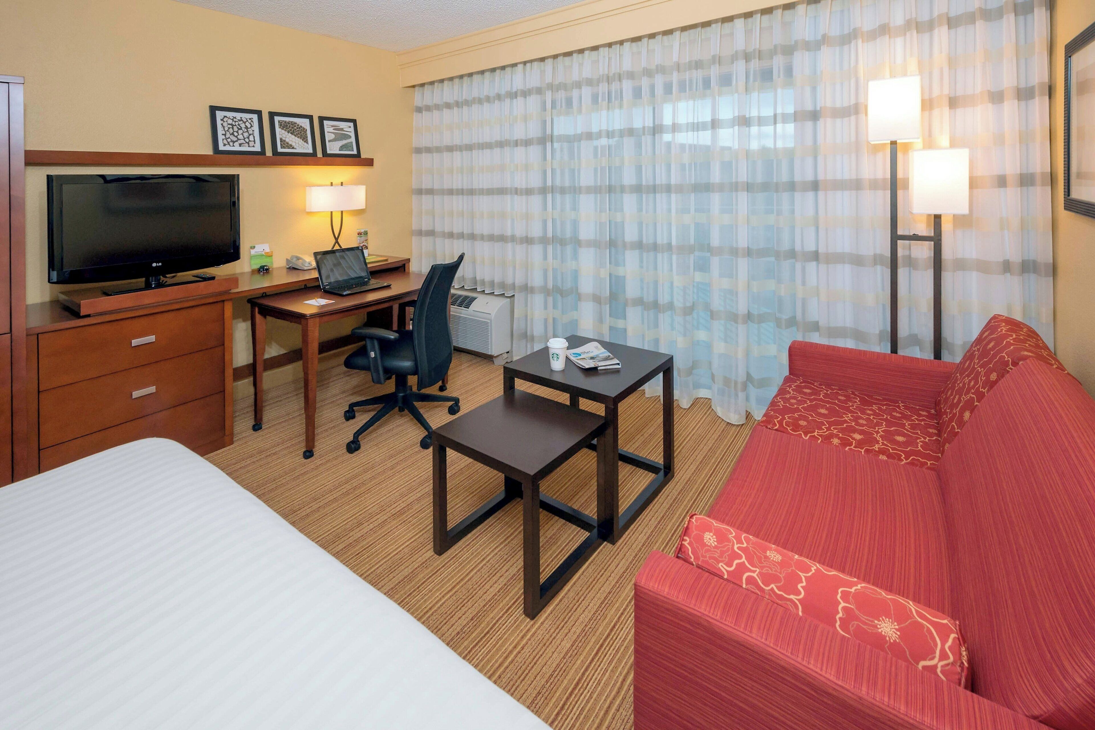 Ein Hotelzimmer mit Bett, Schreibtisch mit Computer, Fernseher, rotem Sofa und einem Fenster mit Vorhängen.