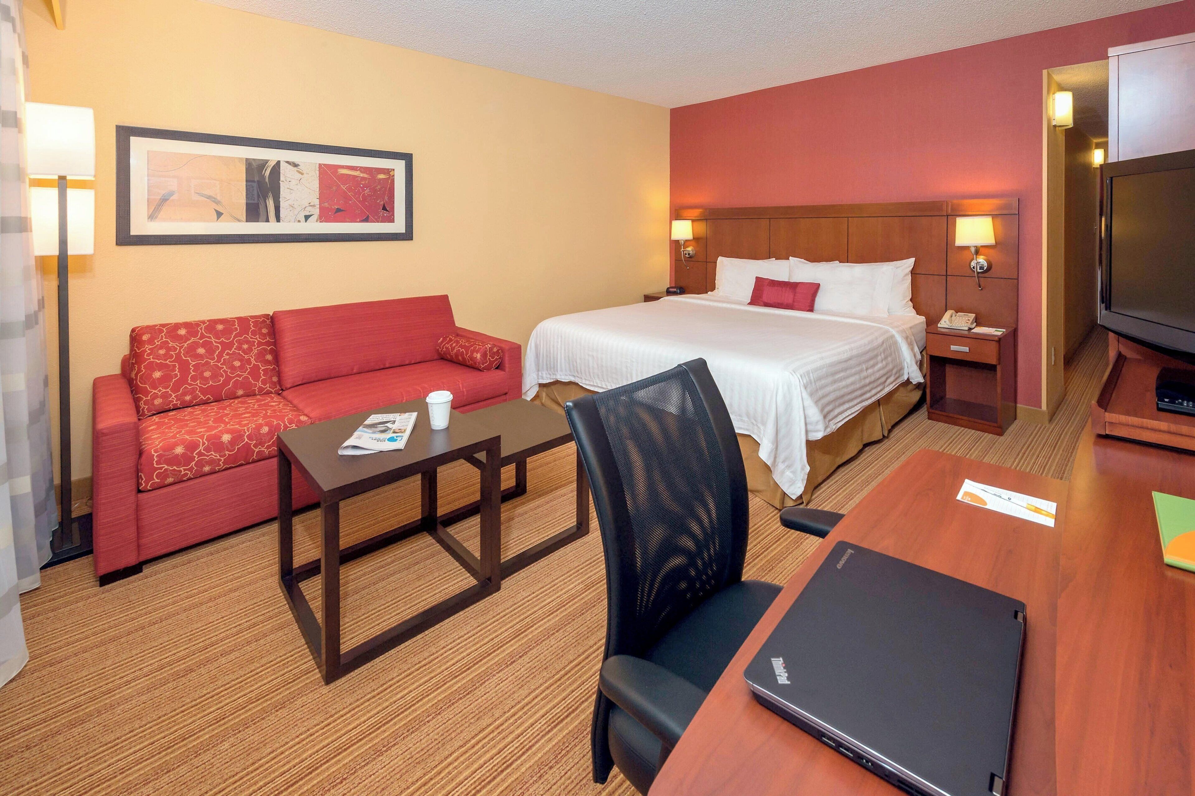 A hotel room with a bed, a red sofa, a wooden table, a laptop, and a television.