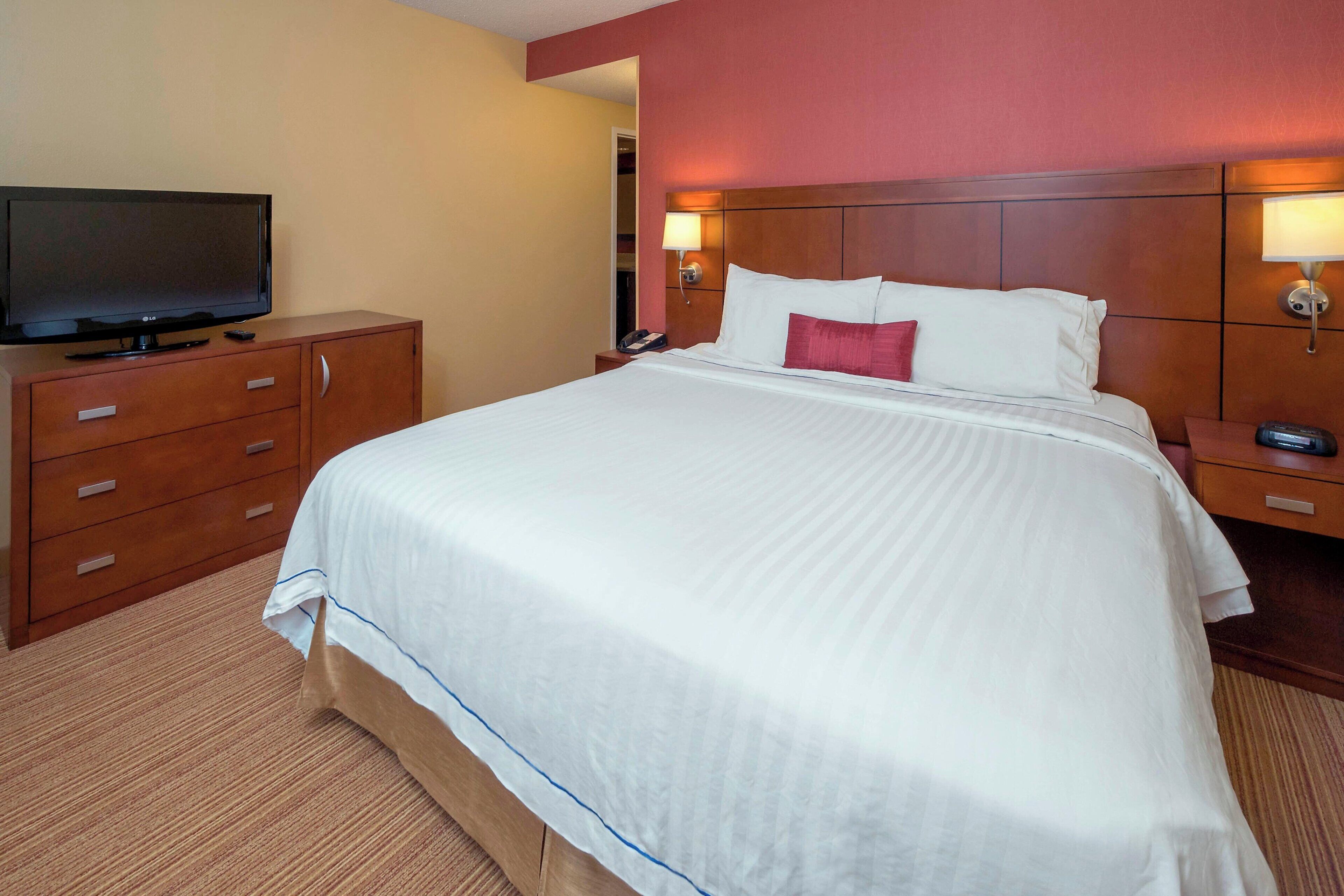 A hotel room with a large bed, wooden headboard, bedside tables with lamps, a flat-screen TV on a wooden cabinet, and a carpeted floor.