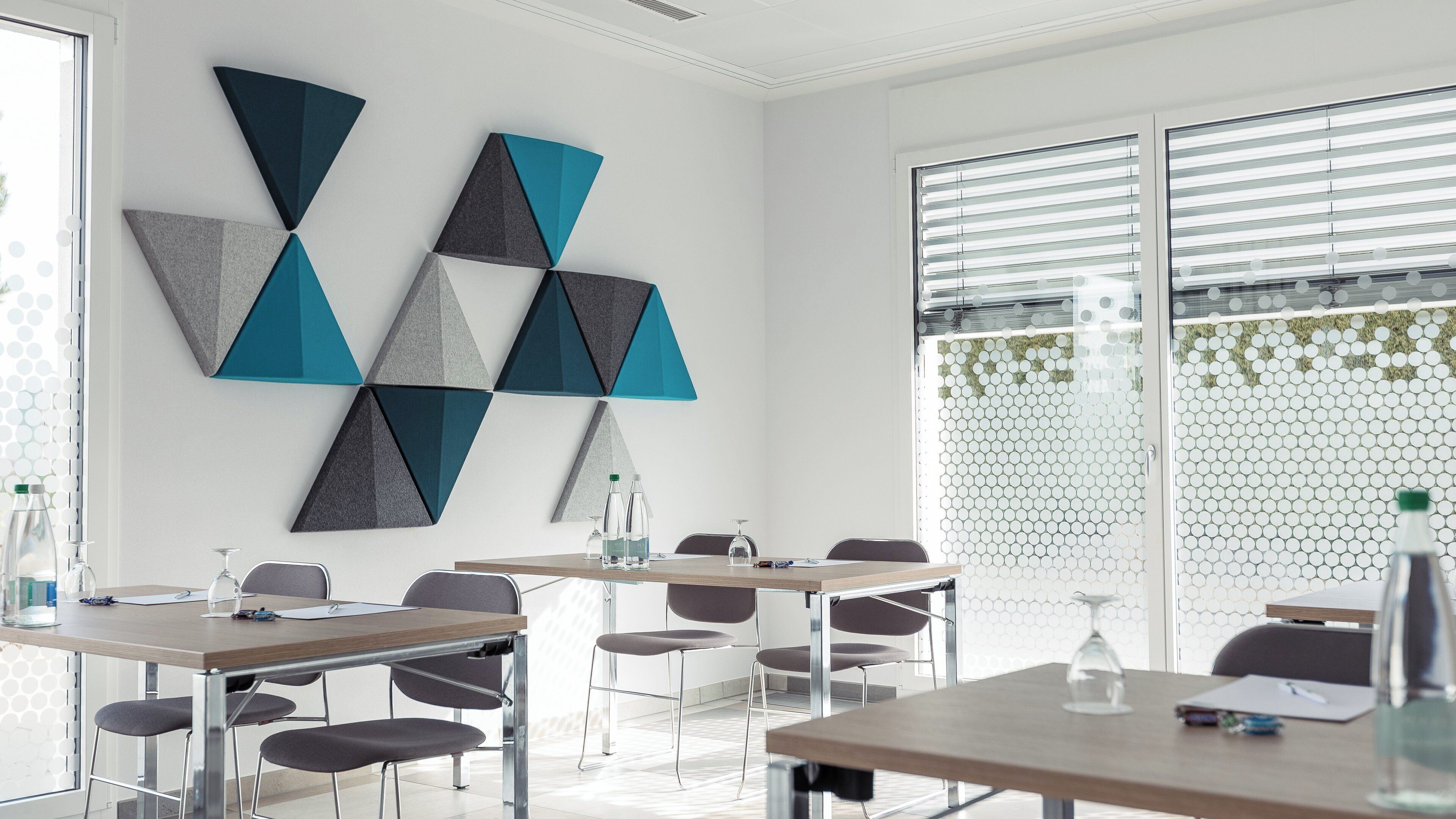 Una sala de conferencias moderna con arte geométrico en la pared, un ventanal con persianas y una mesa de reuniones con sillas.