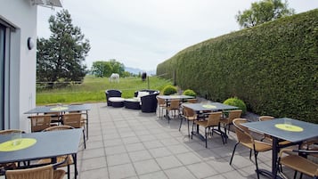 An outdoor patio with tables and chairs, a hedge, and a horse in the background.
