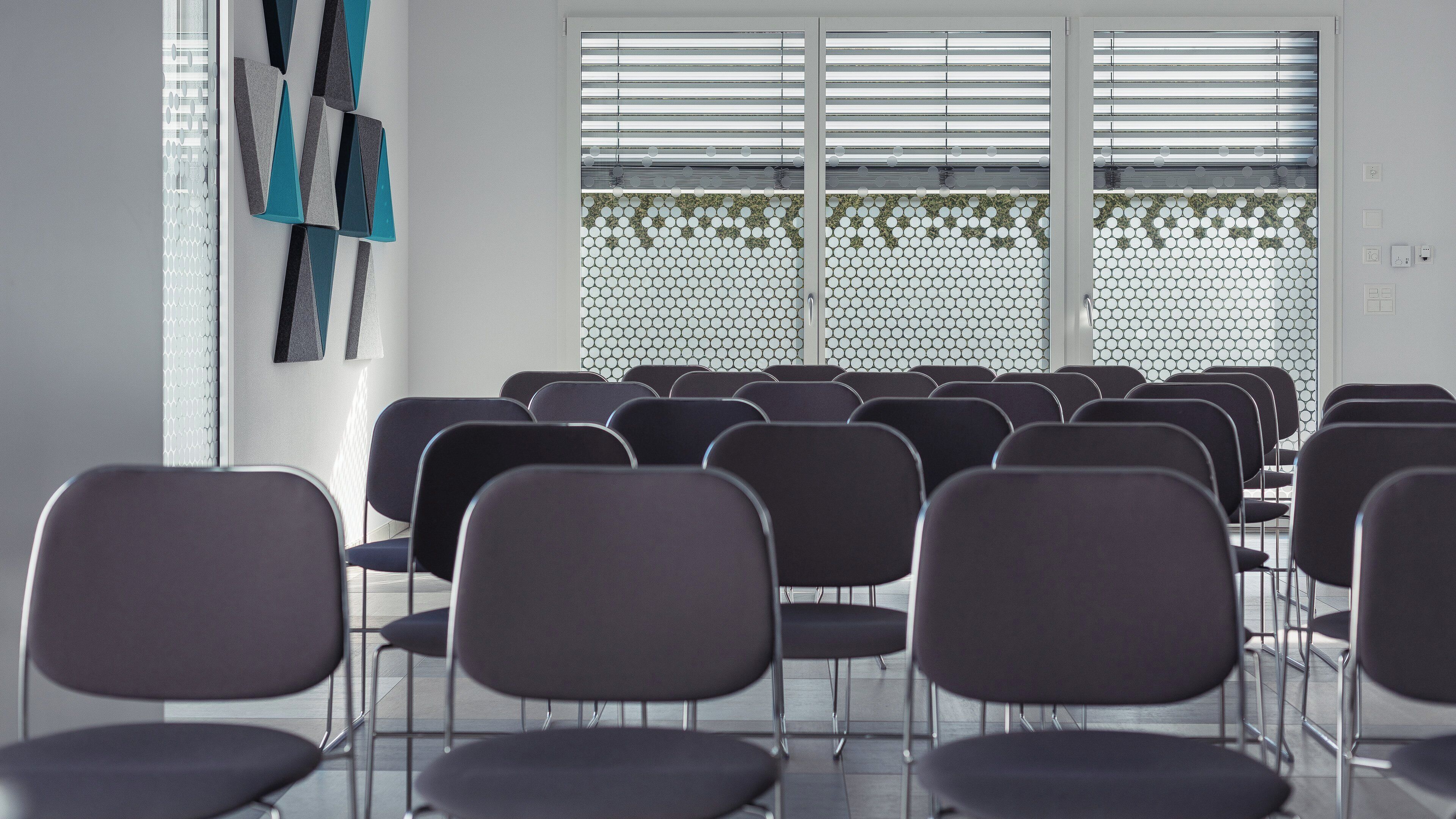 A room with rows of chairs facing a wall with a geometric pattern and a window with horizontal blinds.
