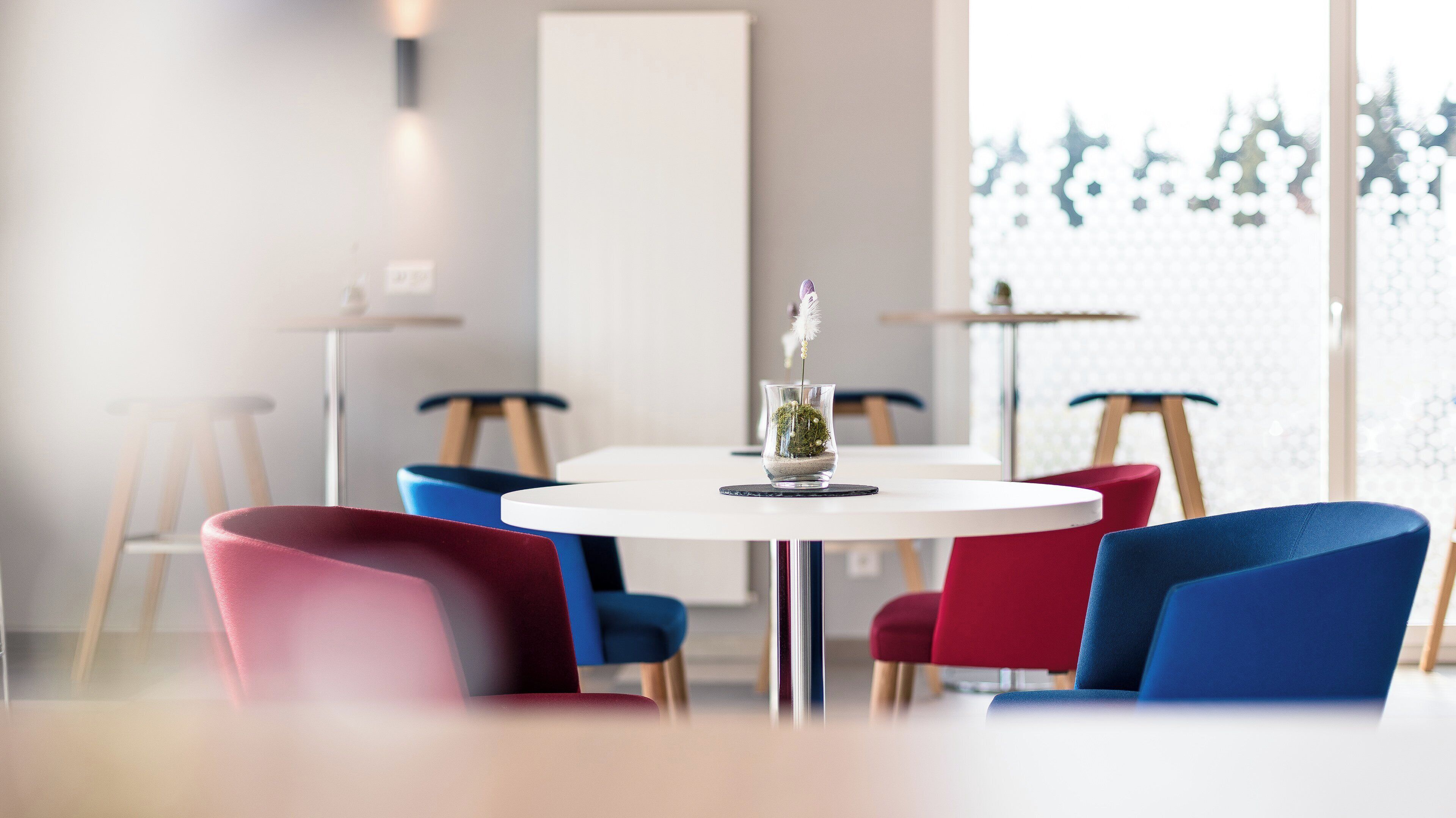 Een moderne eethoek met ronde tafels en stoelen, een decoratieve vaas met plant en uitzicht op bomen door een raam.