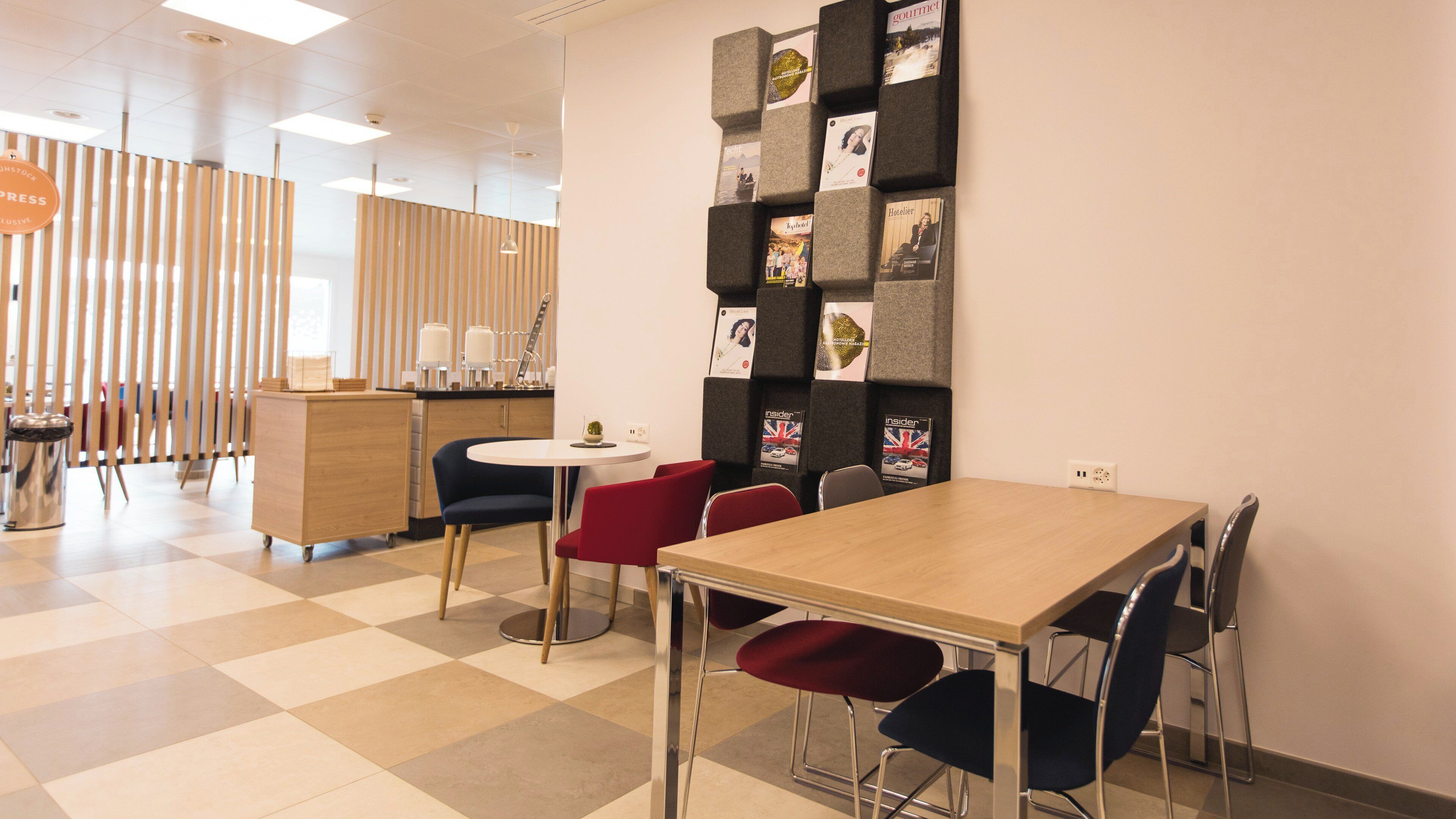 Un espacio de oficina moderno con una mesa de madera, sillas negras y rojas, una mesita redonda y un revistero de pared.