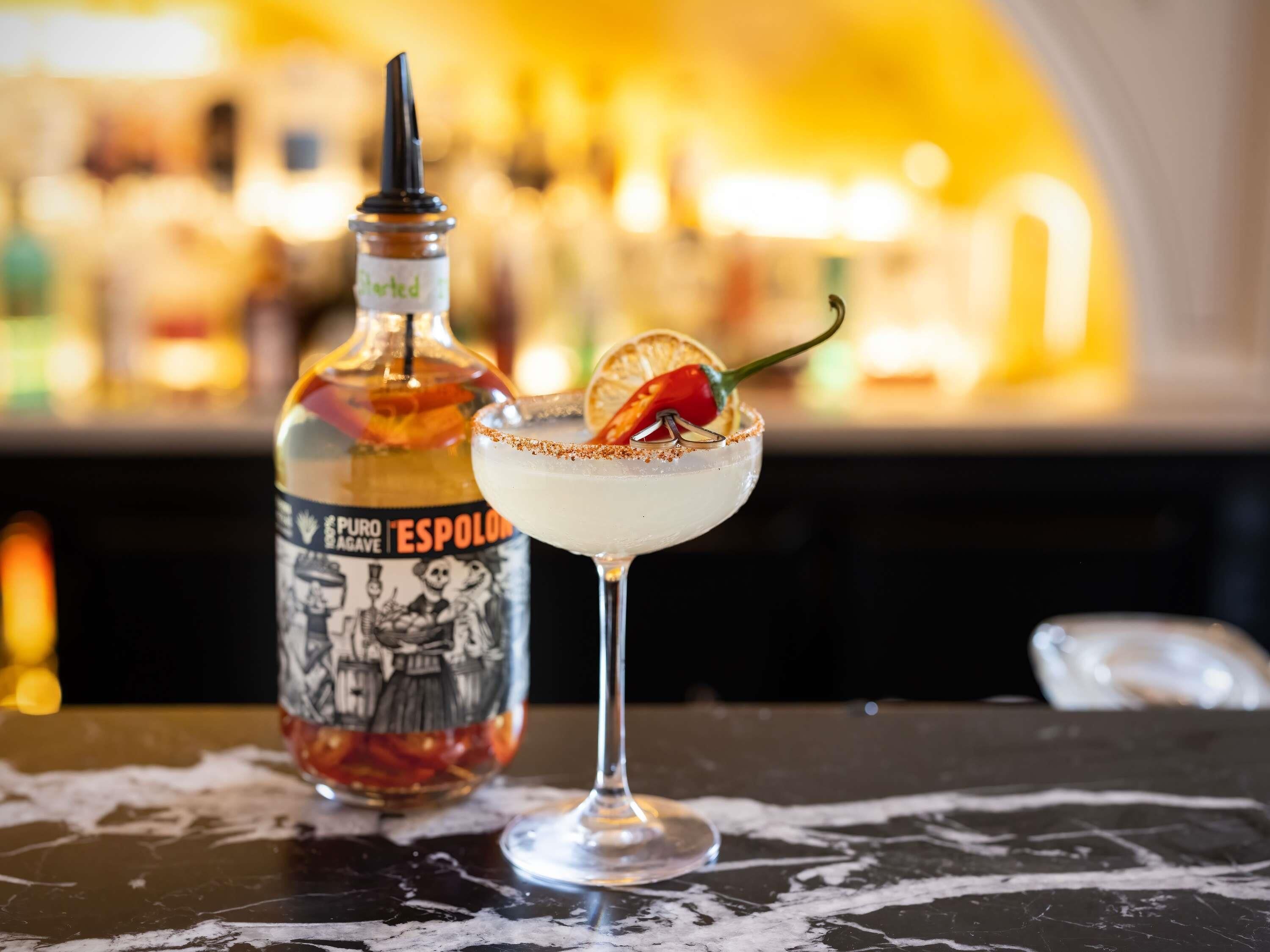 A bar setting with a bottle labeled 'Puro de Agave' and a margarita glass with a garnish.