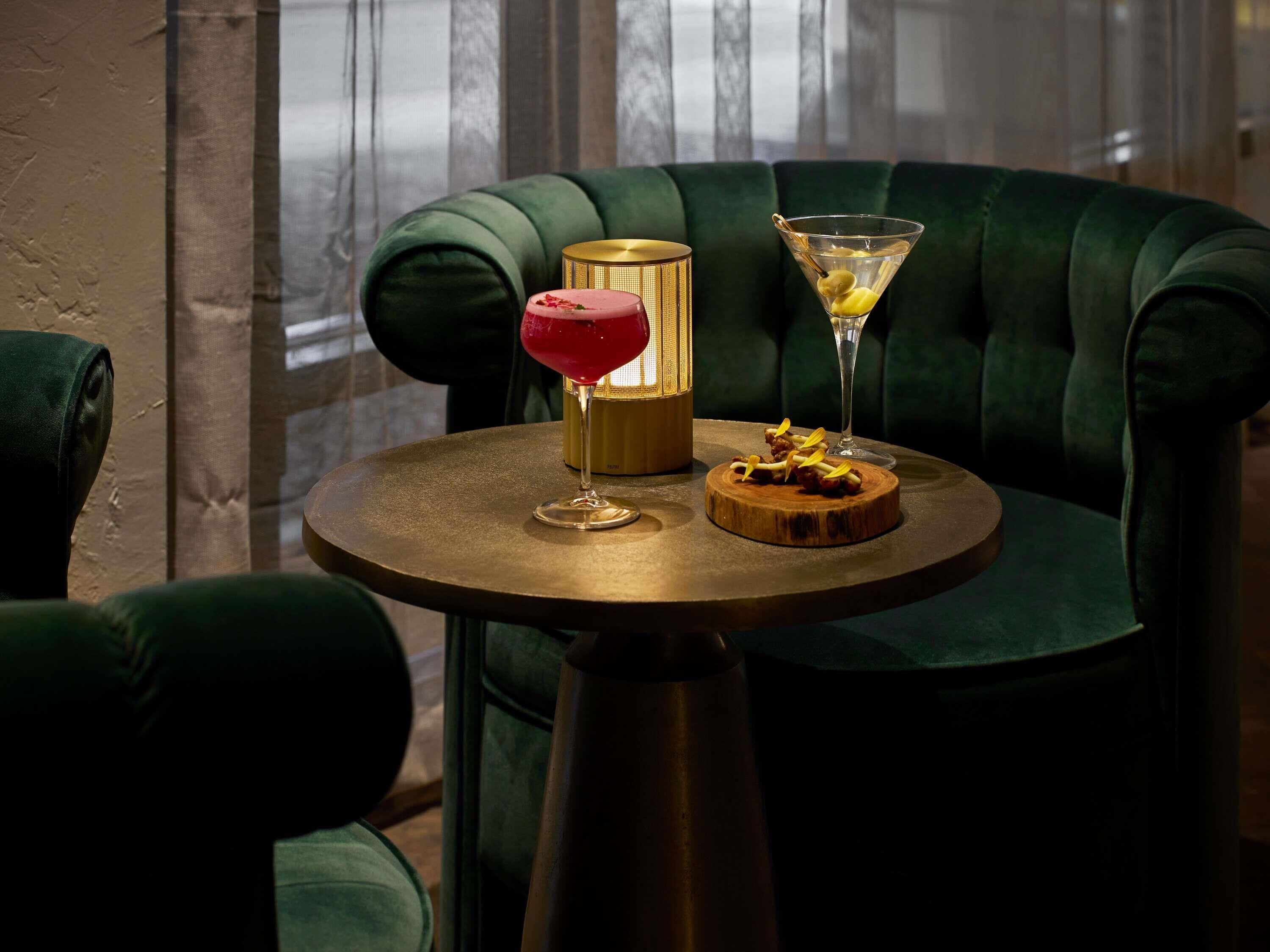 Una mesa redonda con una copa de martini y un cóctel, una tabla de madera con comida y una lámpara con base dorada.