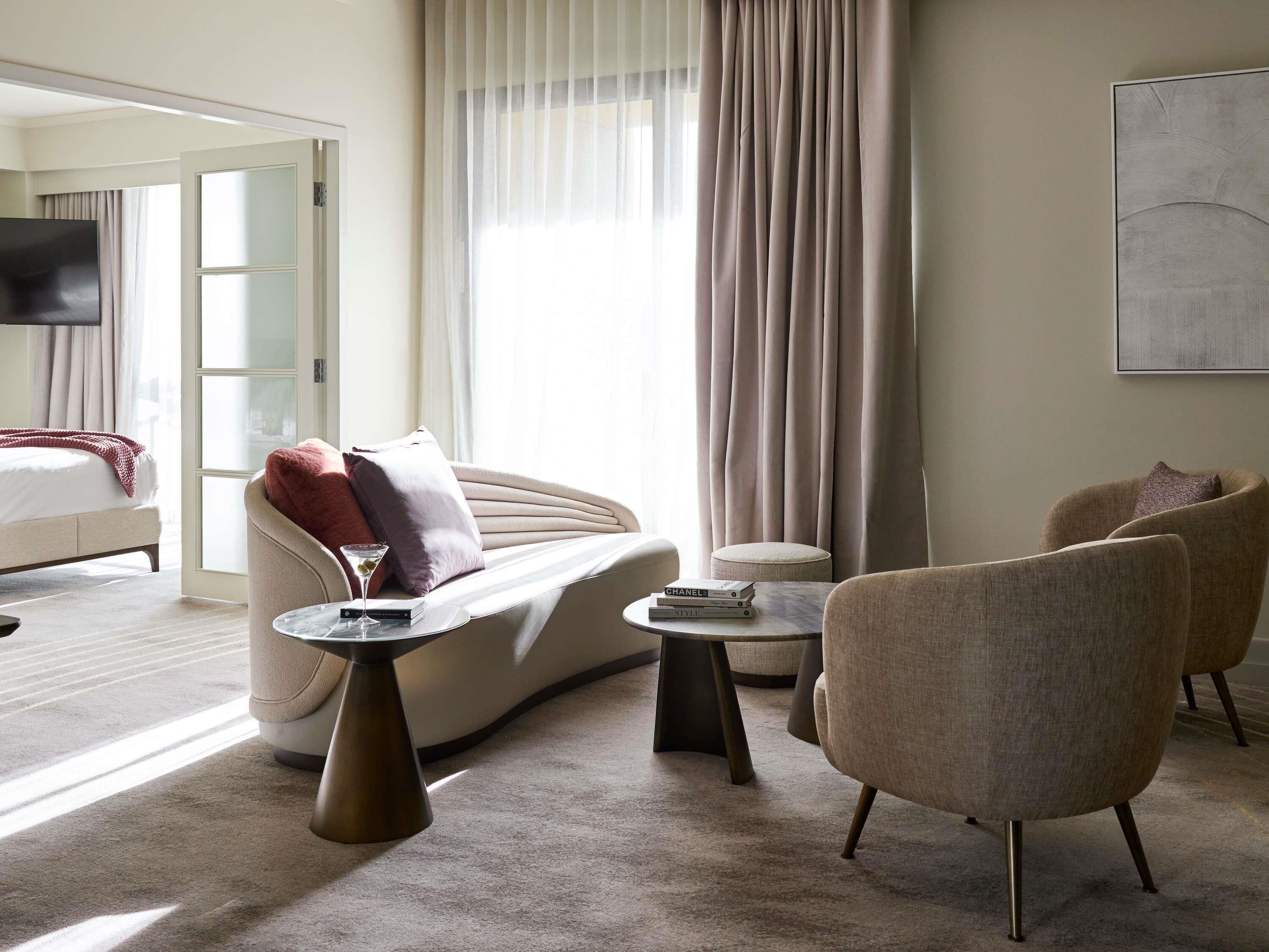 A modern hotel room with a sofa, armchairs, a coffee table, and a TV.