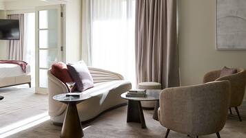 A modern hotel room with a sofa, armchairs, a coffee table, and a TV.