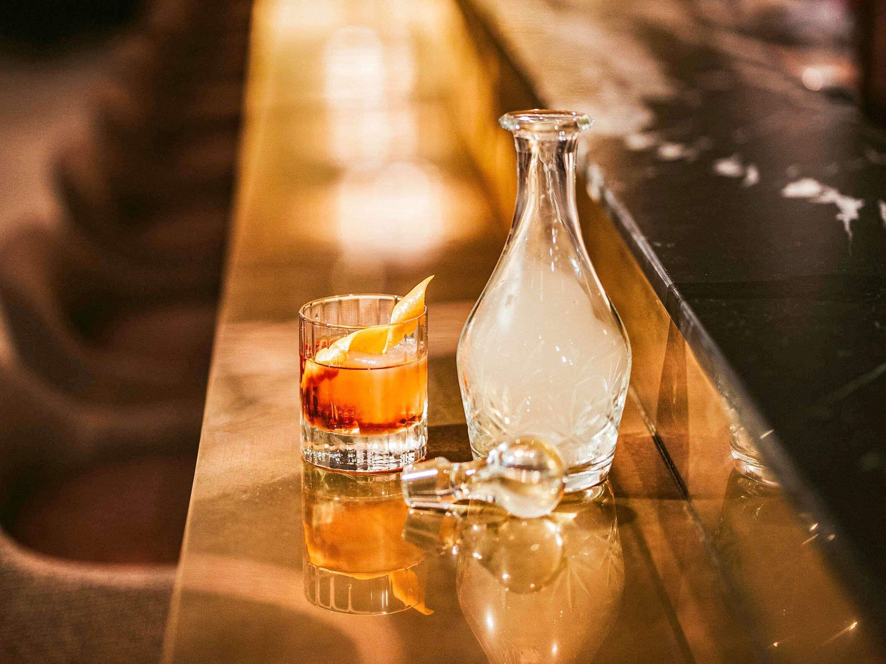 Un vaso de whisky con una cáscara de naranja como adorno sobre una barra reflectante, junto a una botella transparente.