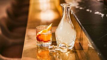 Un vaso de whisky con una cáscara de naranja como adorno sobre una barra reflectante, junto a una botella transparente.