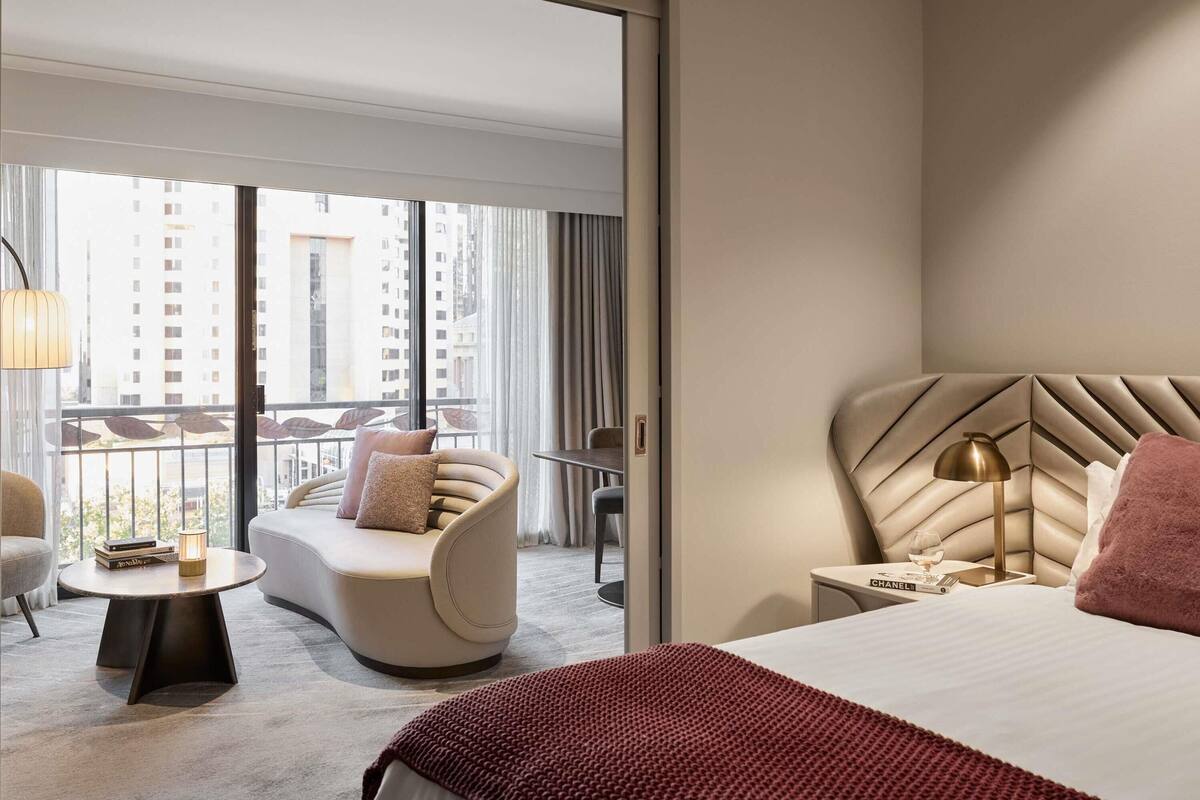 A hotel room with a bed, a sofa, a small table, a lamp, and a view of the city through large windows.