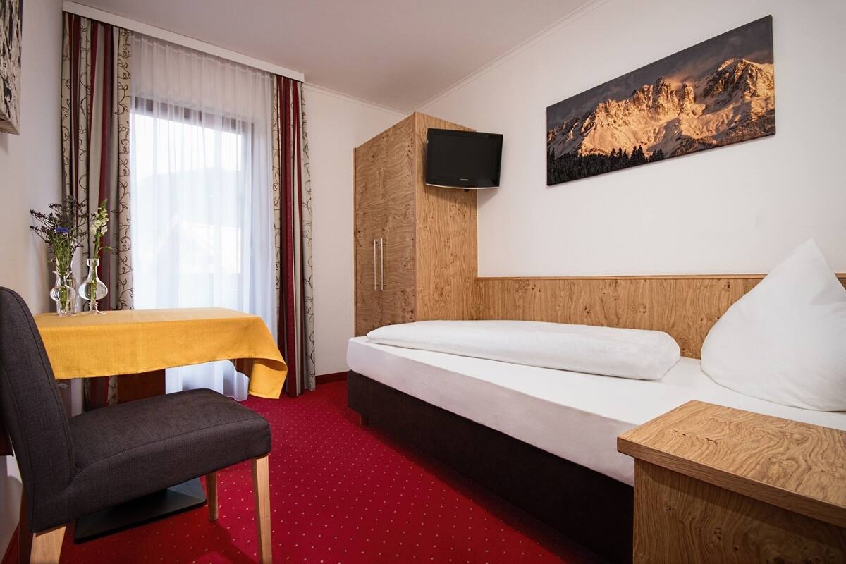 Une chambre d’hôtel avec un lit, une tête de lit en bois, un téléviseur fixé au mur, une petite table recouverte d’une nappe jaune, une chaise et une moquette rouge.