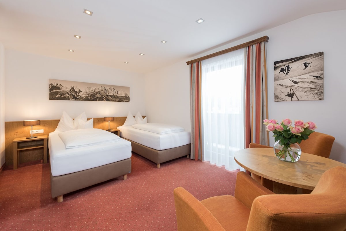 Une chambre d’hôtel avec deux lits, un bureau et des chaises. Il y a un tableau au mur et un vase rempli de fleurs sur le bureau.