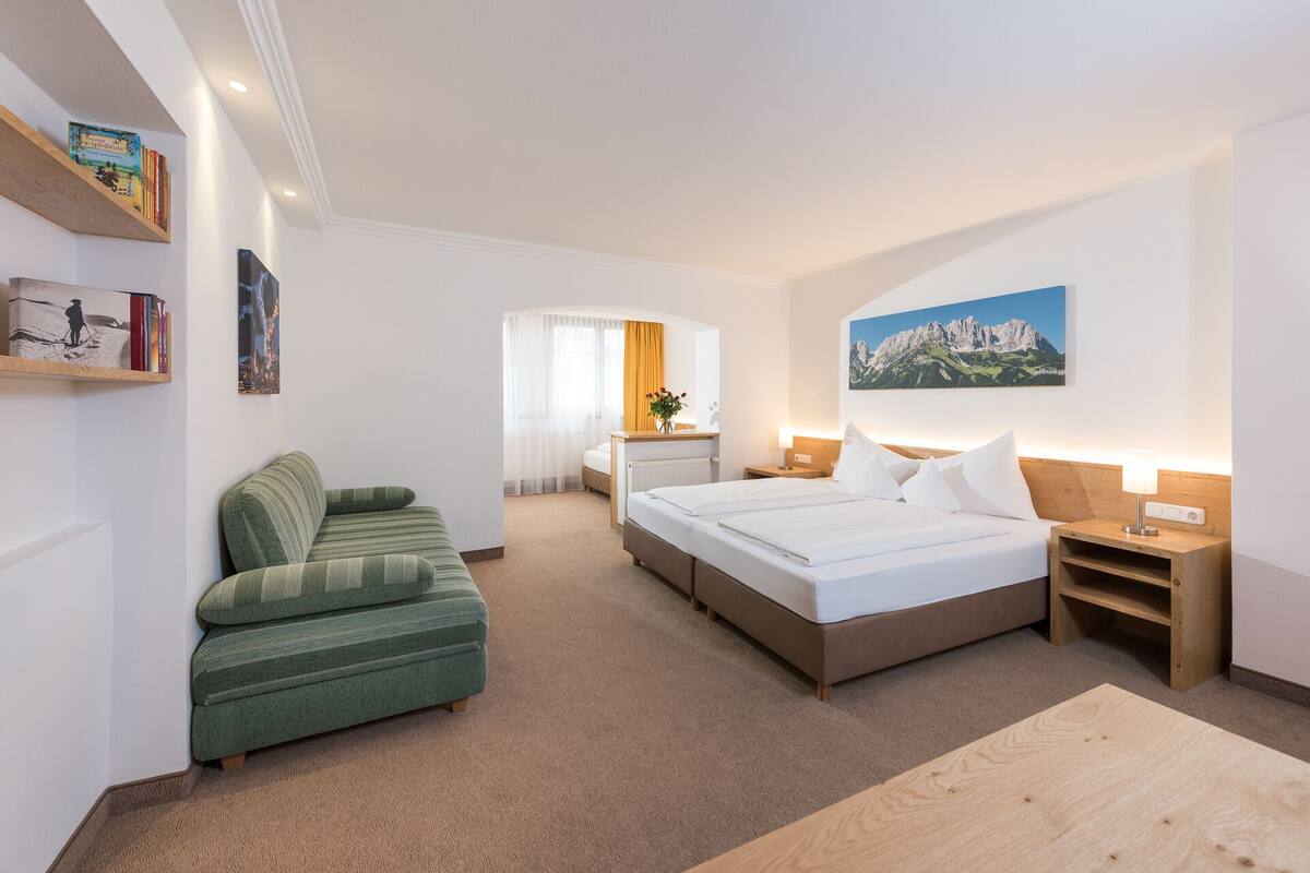 Une chambre d’hôtel avec un lit, un canapé vert, une table de chevet en bois, une table en bois, un tableau et une bibliothèque contenant des livres et une photographie encadrée.