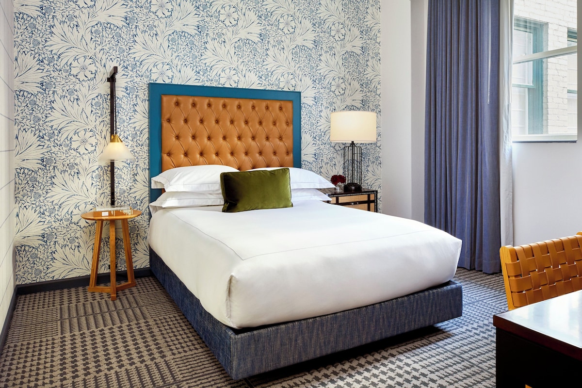 A hotel room with a bed, a nightstand, a lamp, a chair, and patterned wallpaper.