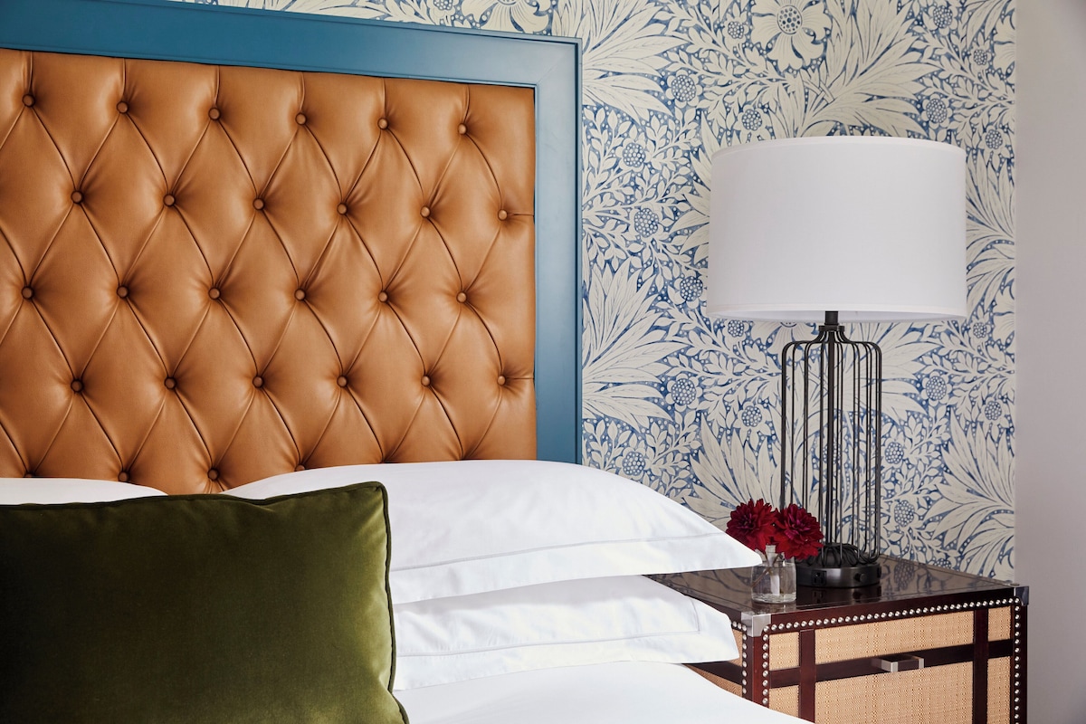 A bed with a tufted headboard, white bedding, a green pillow, and a nightstand with a lamp and flowers.