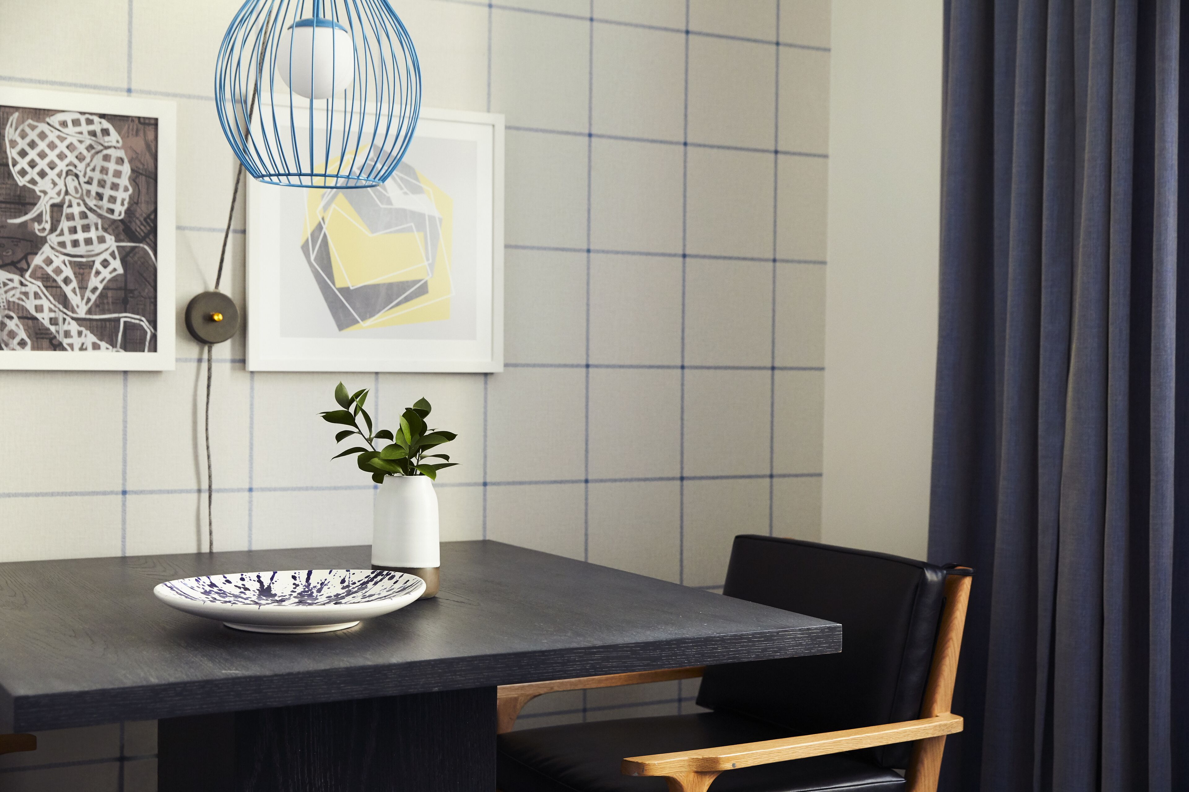 A dining area with a dark table, a white vase with a plant, and framed artwork on the wall.