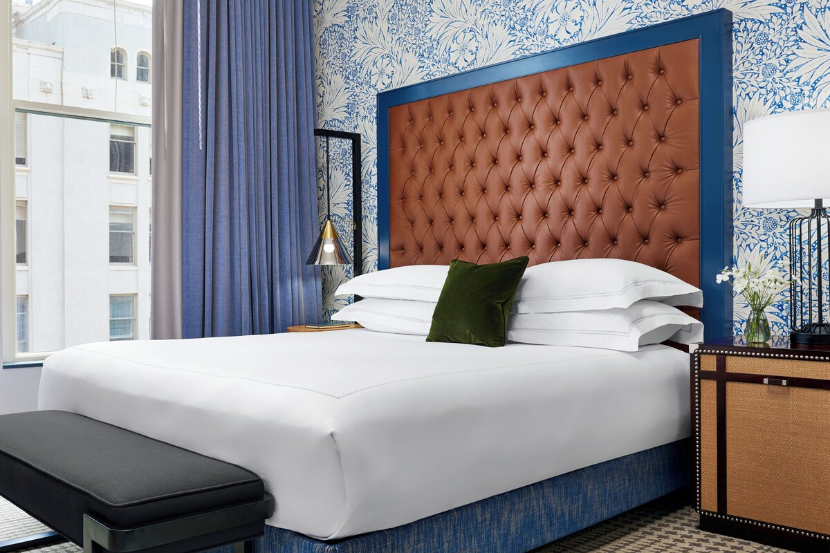A hotel room with a large bed, a bench, a nightstand, and a view of the city.