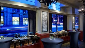 A bar area with a view of a city street at night, featuring a bar counter with bottles and glasses, and a chandelier.