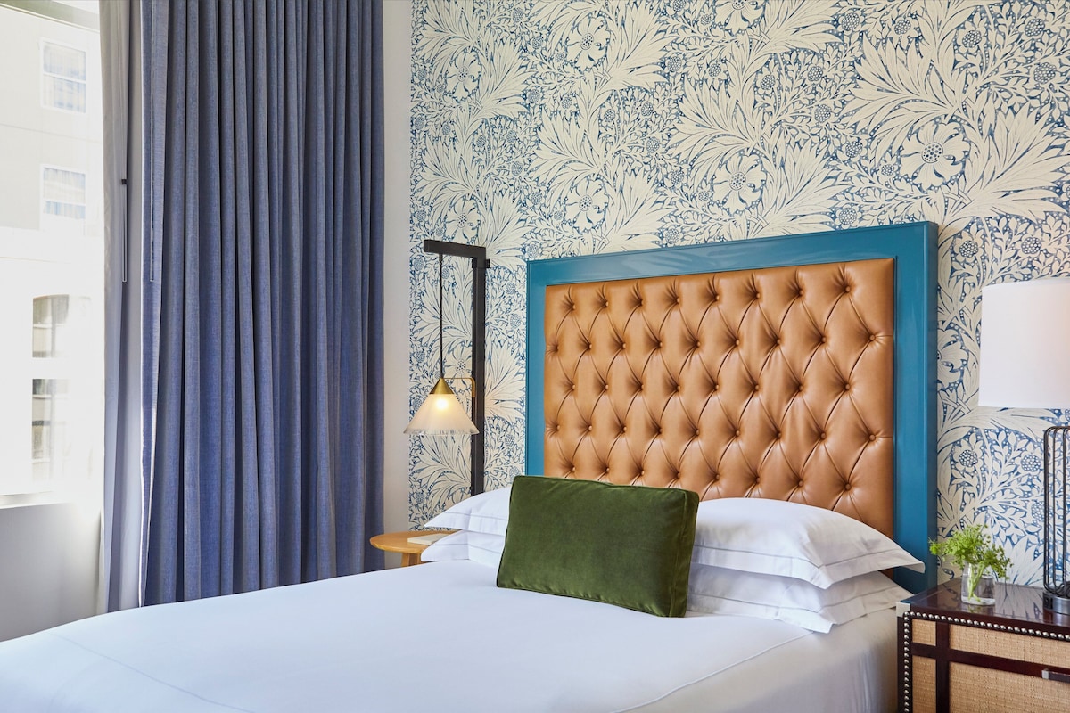 A hotel room with a large bed, a headboard, a nightstand, a lamp, and a window with curtains.