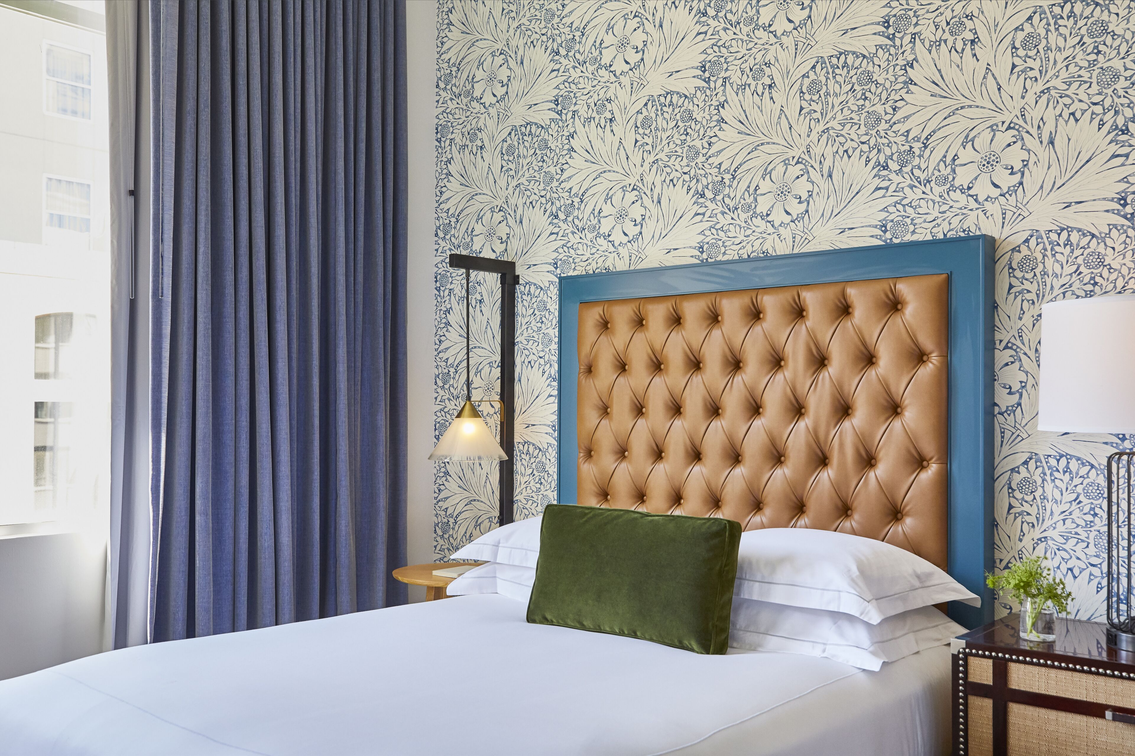 Une chambre d’hôtel avec une tête de lit capitonnée, un lit recouvert de draps blancs, un oreiller vert, une table de chevet avec une lampe et un papier peint à motifs.