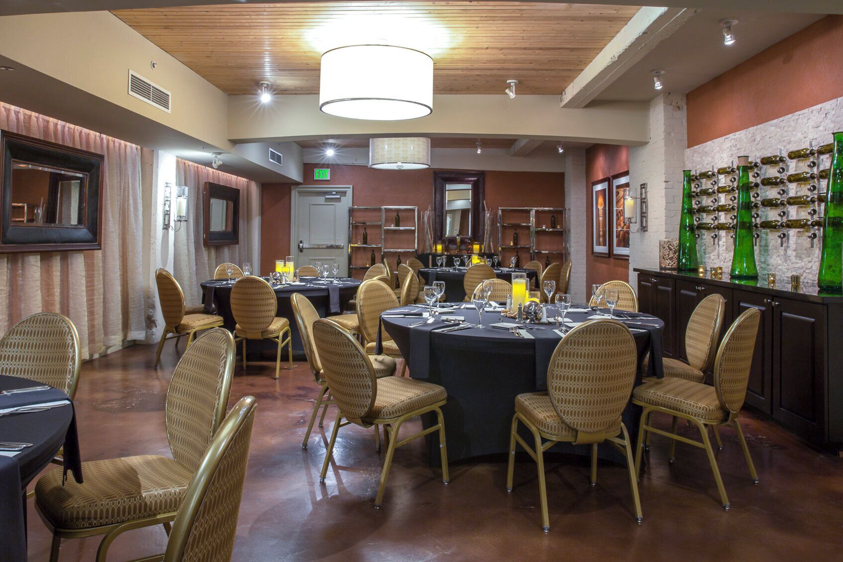 A restaurant with round tables, wicker chairs, and a wall of green bottles.