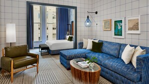 A modern hotel room with a blue sofa, a green armchair, a wooden coffee table, and a bed with a blue curtain.