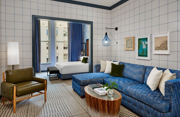 Habitació d'hotel moderna amb un sofà blau, una butaca verda, una taula baixa de fusta i un llit amb cortina blava.