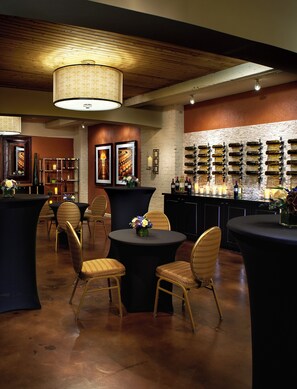 A bar area with round tables, wicker chairs, and a wall of wine bottles.