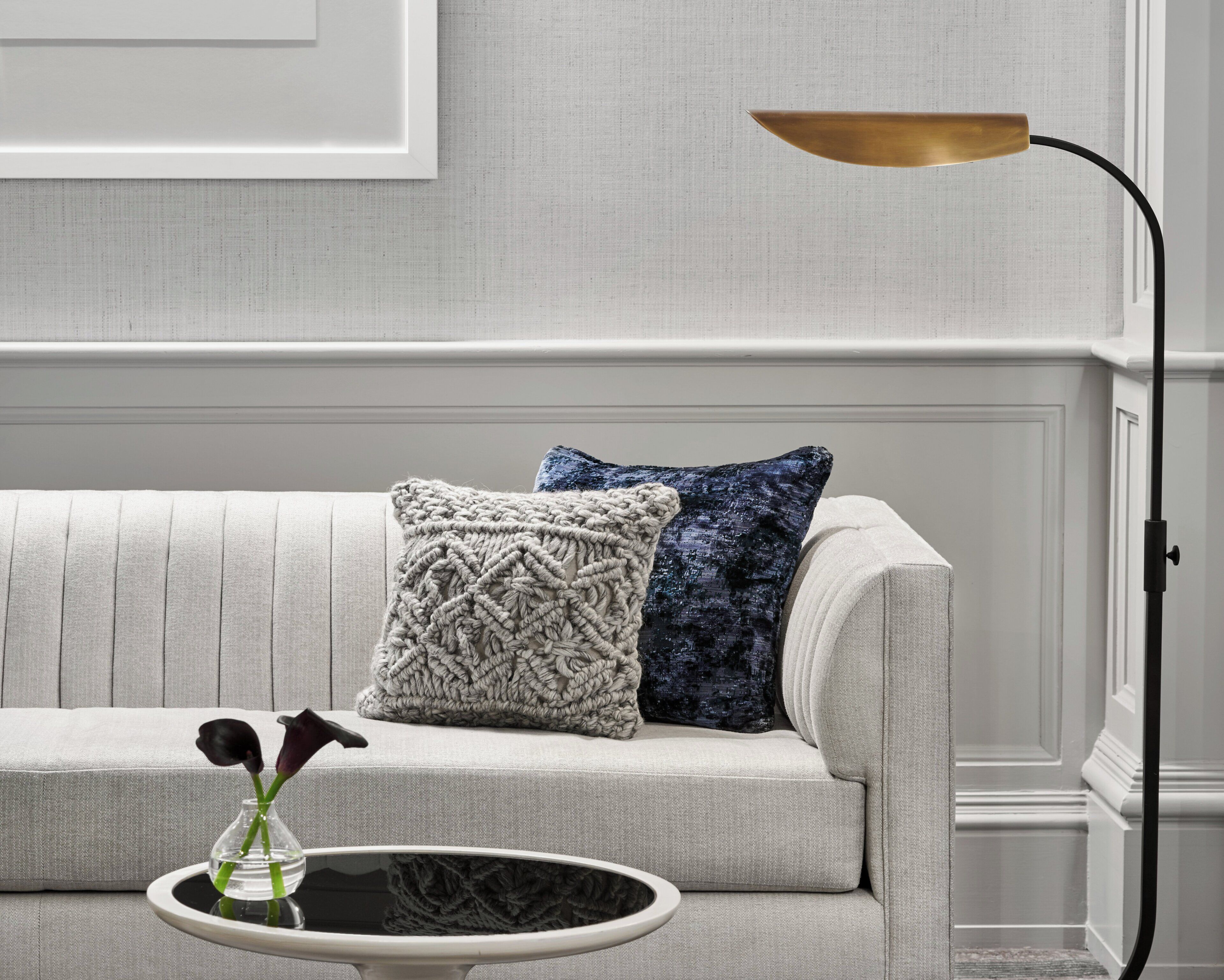 Uma sala de estar moderna com um sofá, dois travesseiros decorativos, uma pequena mesa lateral com um vaso e um abajur.
