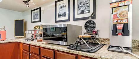 A kitchen with a microwave, coffee machine, and a waffle maker. There are framed pictures on the wall and a speaker on the left.
