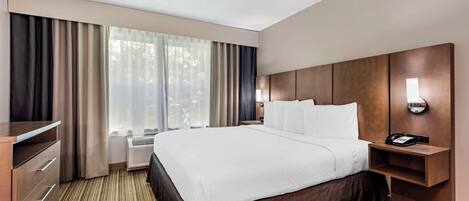 A hotel room with a large bed, a desk, a chair, a nightstand with a lamp, and a window with curtains.