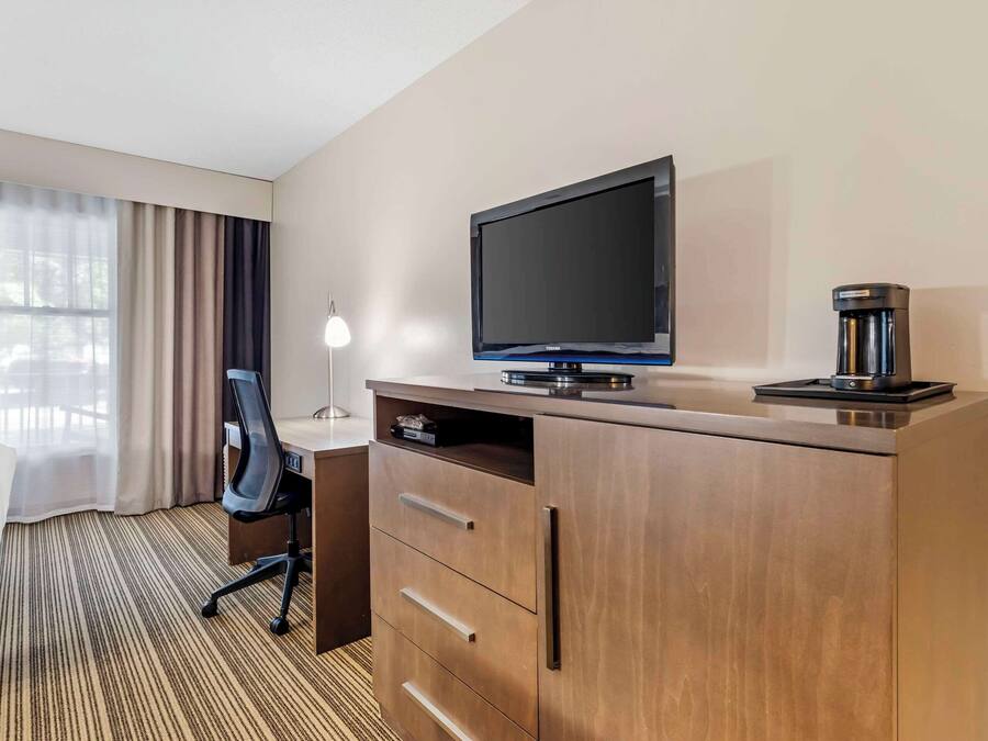 A hotel room with a wooden desk, a flat-screen TV, a coffee maker, a lamp, and a bed with a striped carpet.