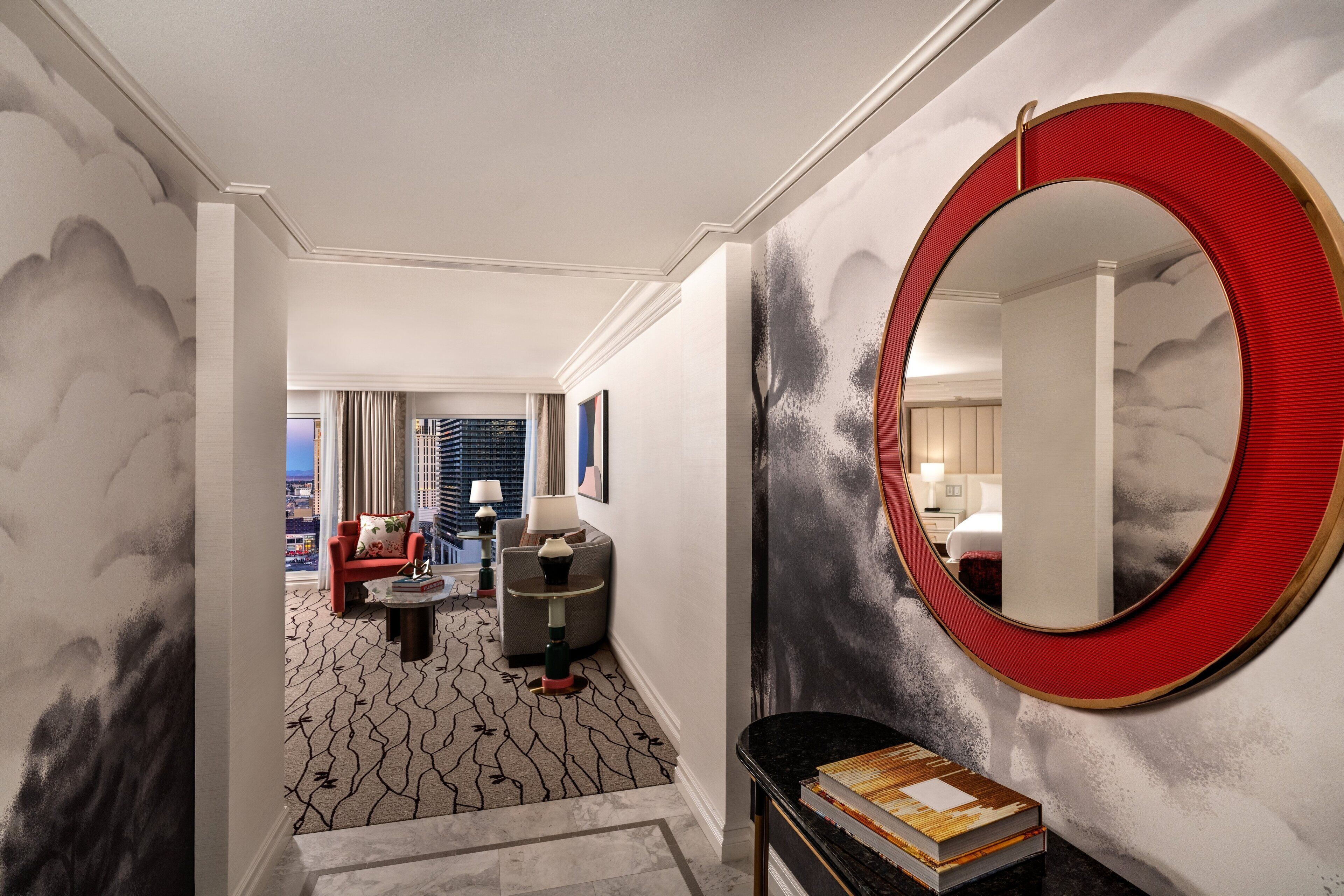 A hotel lobby with a modern design, featuring a round mirror, a seating area with a sofa and armchairs, and a view of the city through large windows.