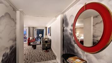 A hotel lobby with a modern design, featuring a round mirror, a seating area with a sofa and armchairs, and a view of the city through large windows.