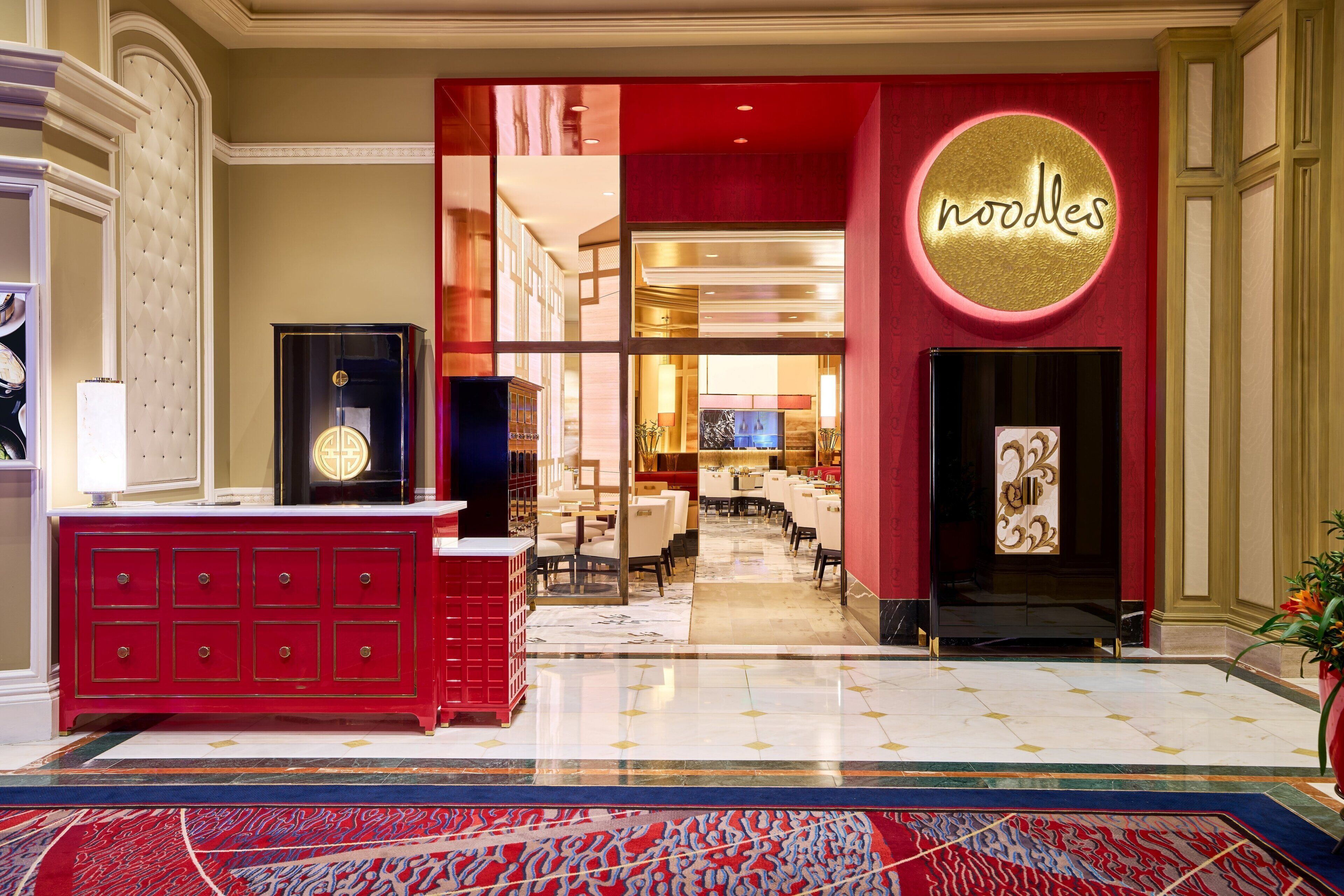 A hotel lobby with a red and gold color scheme, a neon sign reading "noodles", and a reception desk.