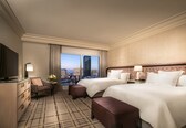 A hotel room with two beds, a TV, a desk, and a view of the cityscape.