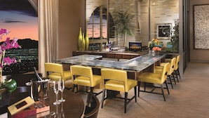 A modern dining area with a long table, yellow chairs, a chandelier, and a cityscape view.