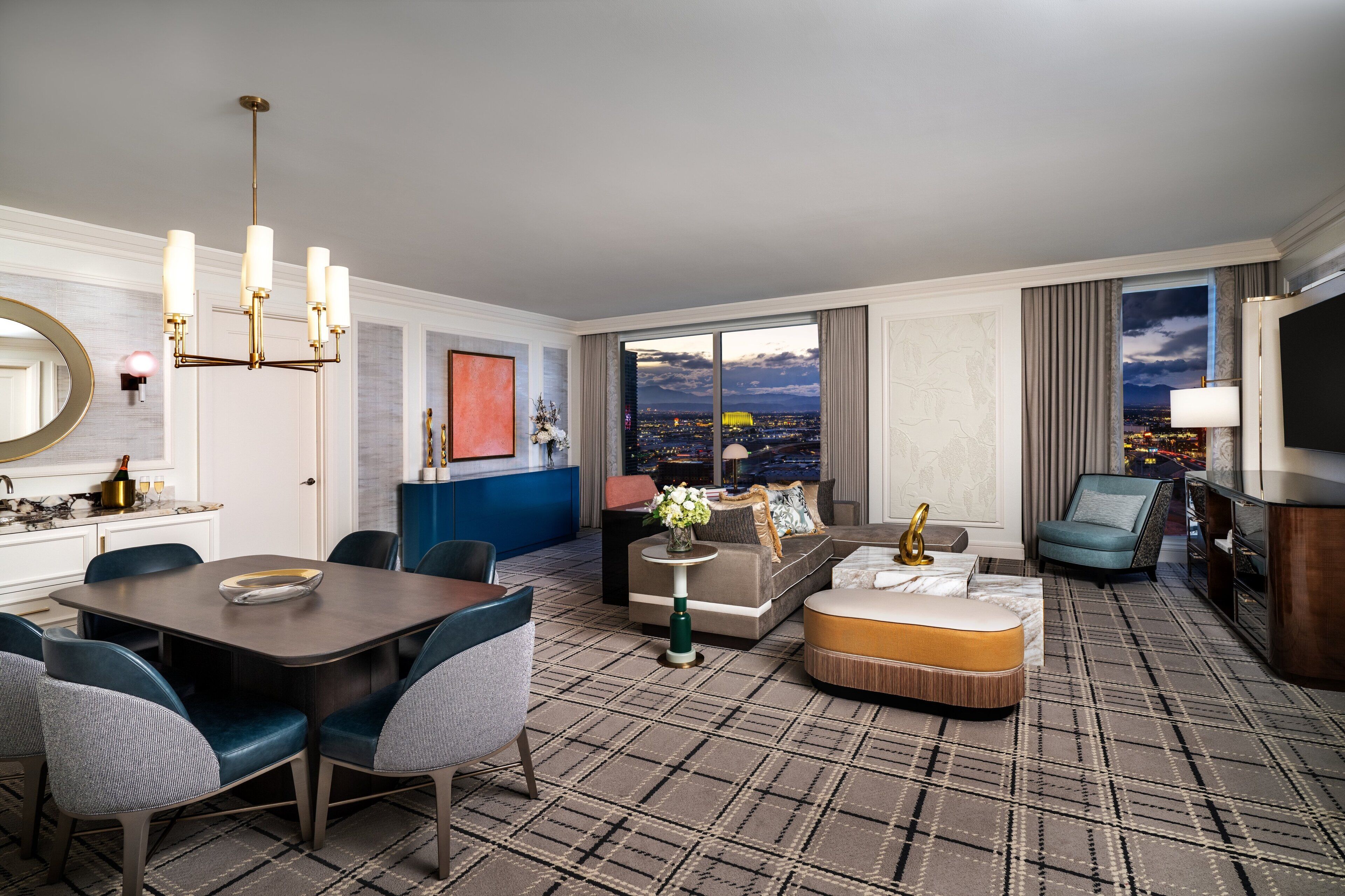 A hotel room with a dining area, a sofa, a TV, and a large window with a city view.