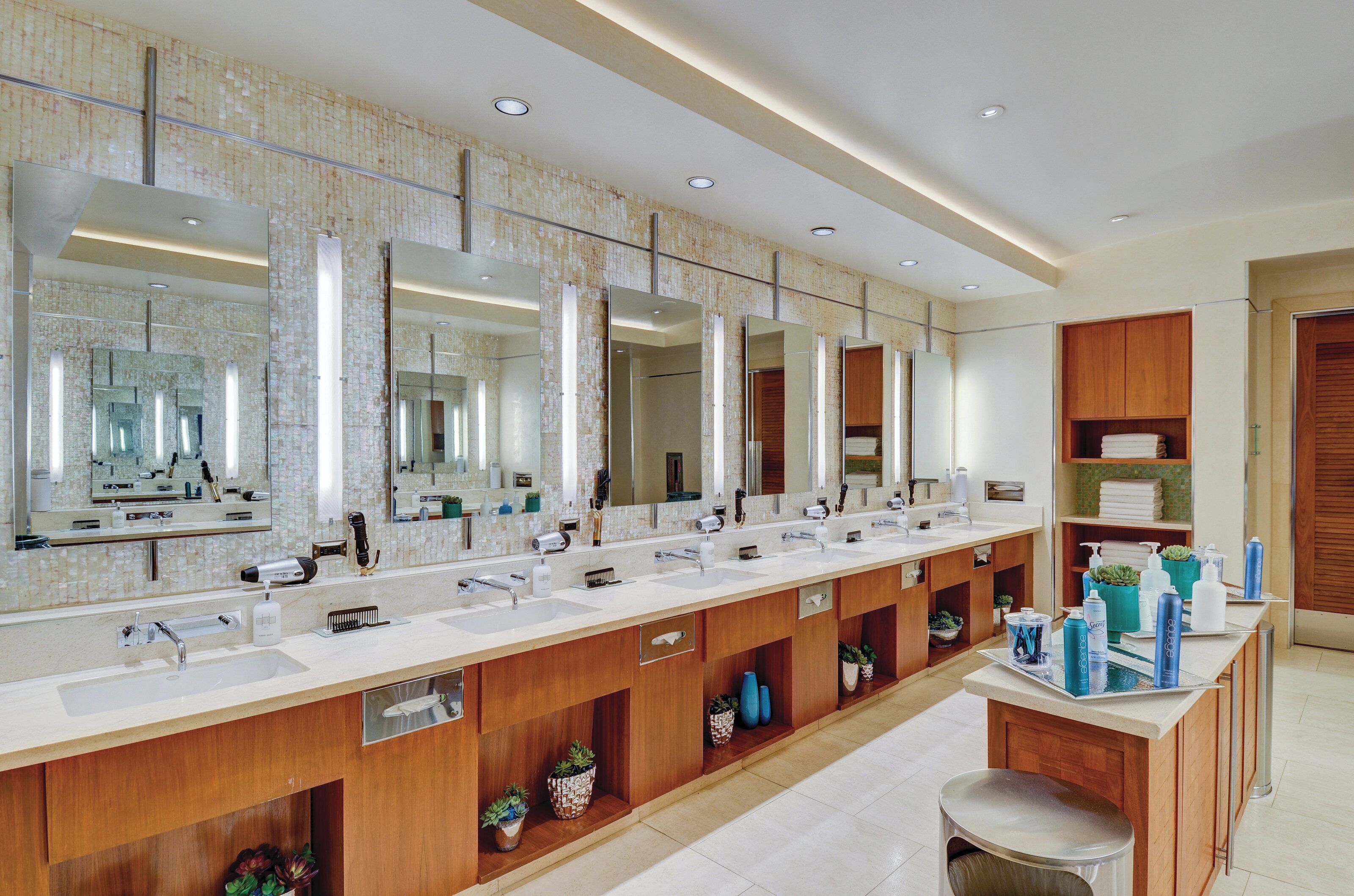Un baño moderno con varios lavamanos, espejos grandes, armarios de madera y plantas en macetas.