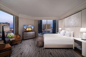 A hotel room with a large bed, a desk with a TV, a chair, a small table with a vase, and a view of the cityscape. - Bellagio (Las Vegas)