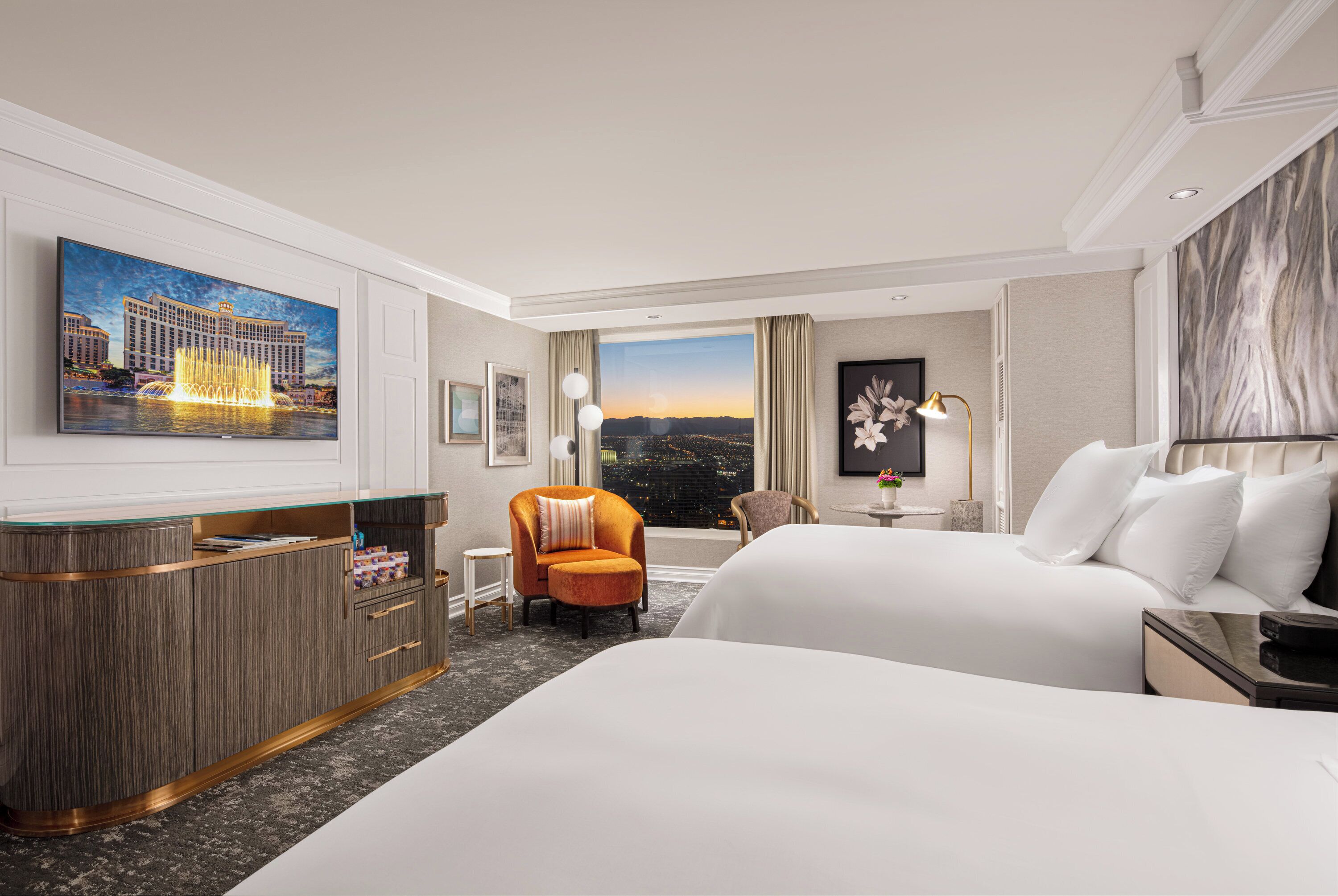 A hotel room with a large bed, a desk with a glass top, an orange armchair, a view of a cityscape, and a painting of a building.