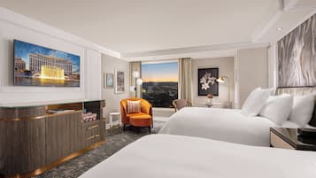 A hotel room with a large bed, a desk with a glass top, an orange armchair, a view of a cityscape, and a painting of a building.
